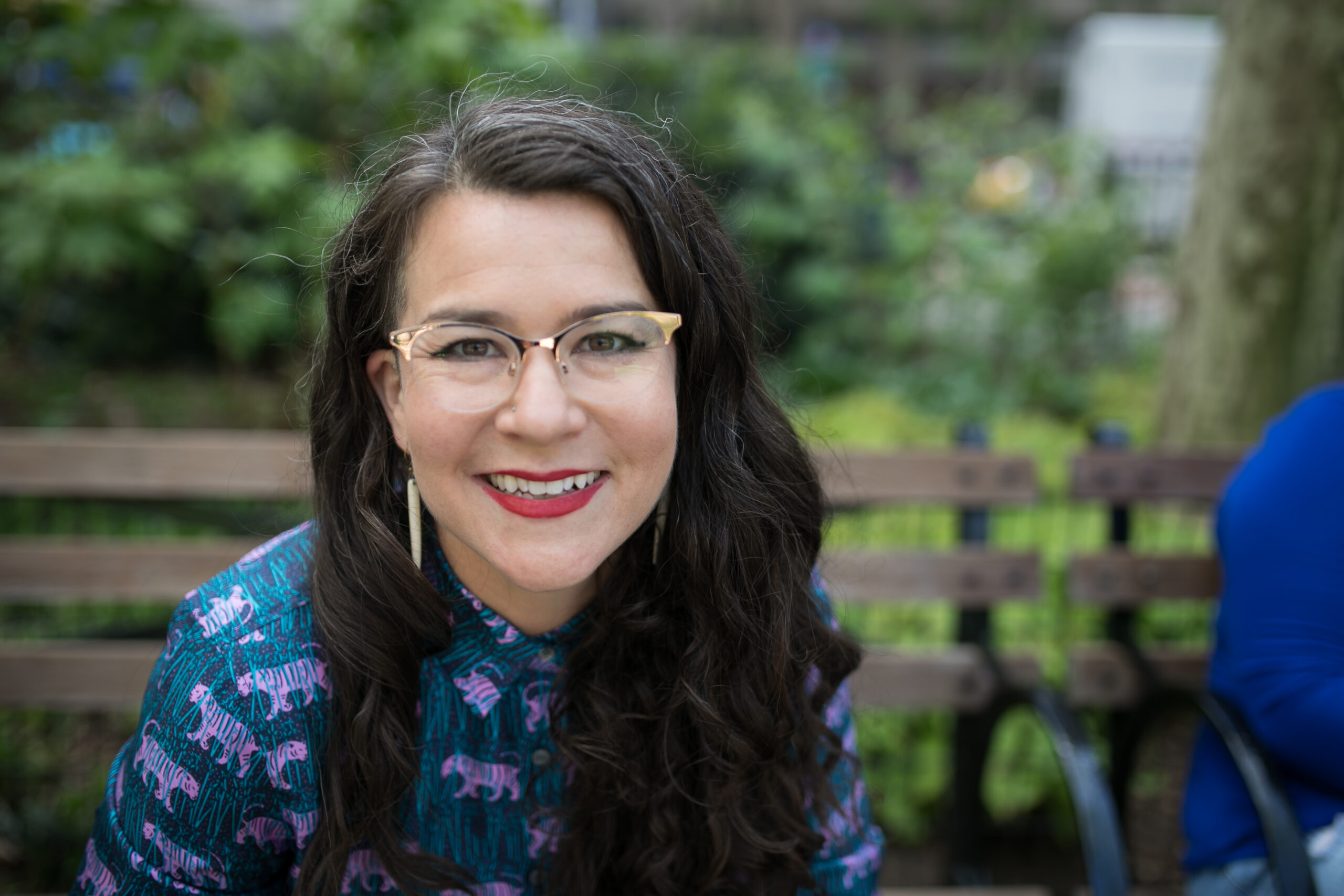 Victoria Albina on bench, gold glasses, looking right at camera outside