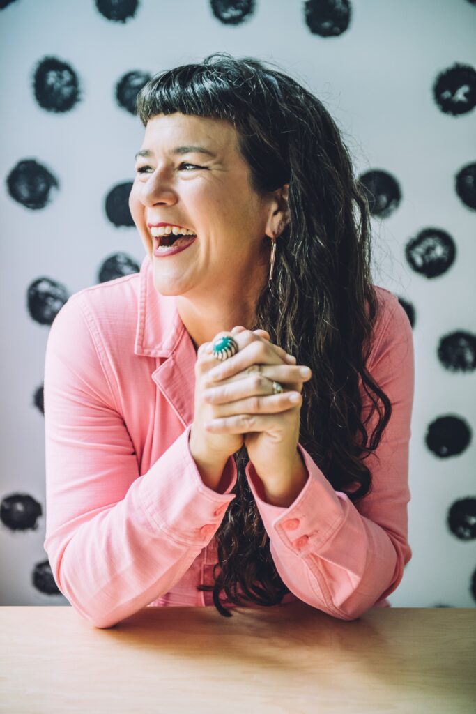 Life coach Victoria Albina in a pink jumpsuit, smiling while sitting at a table